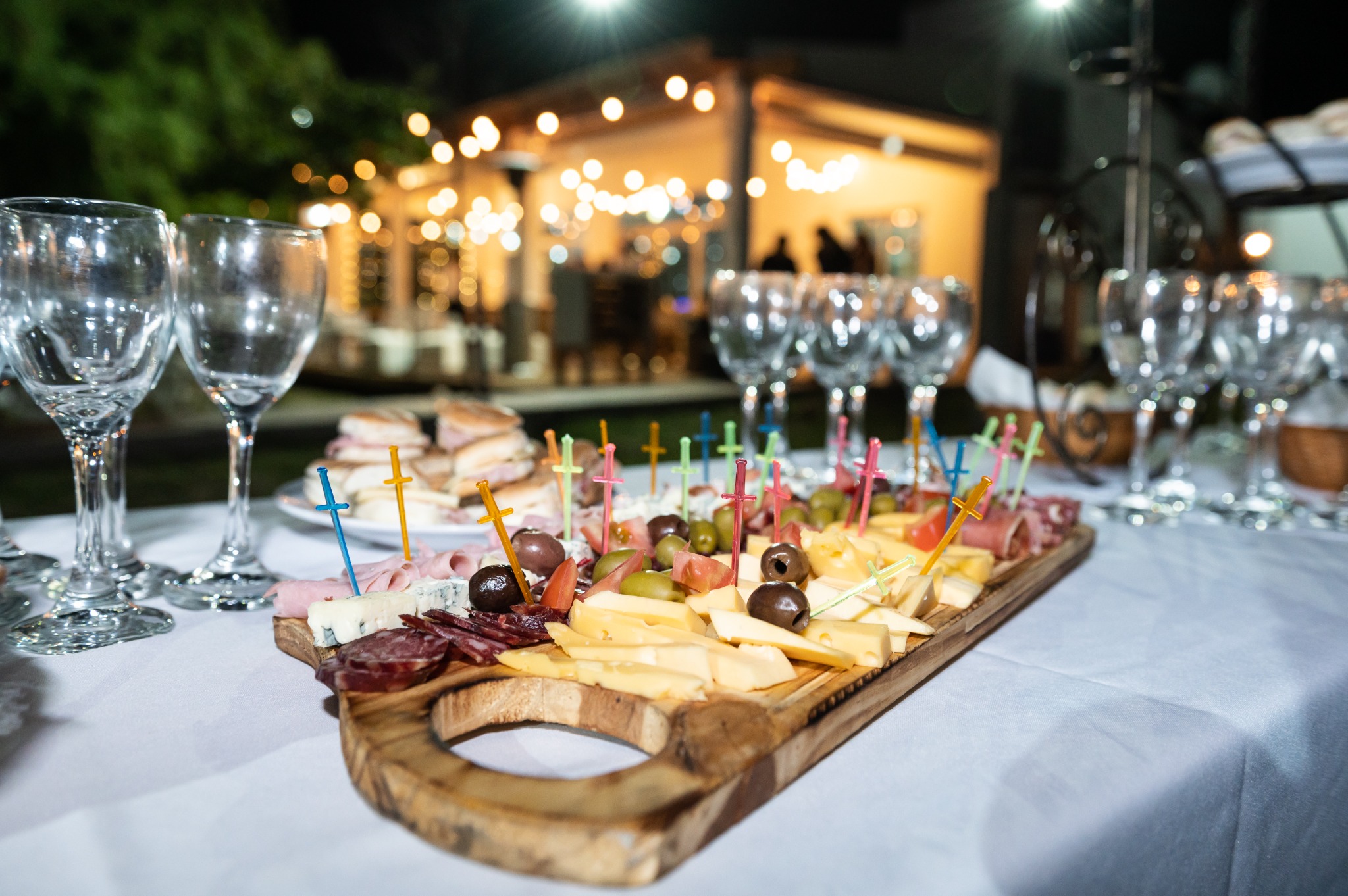 Tabla de quesos, fiambres y aceitunas con copas y la quinta iluminada al fondo